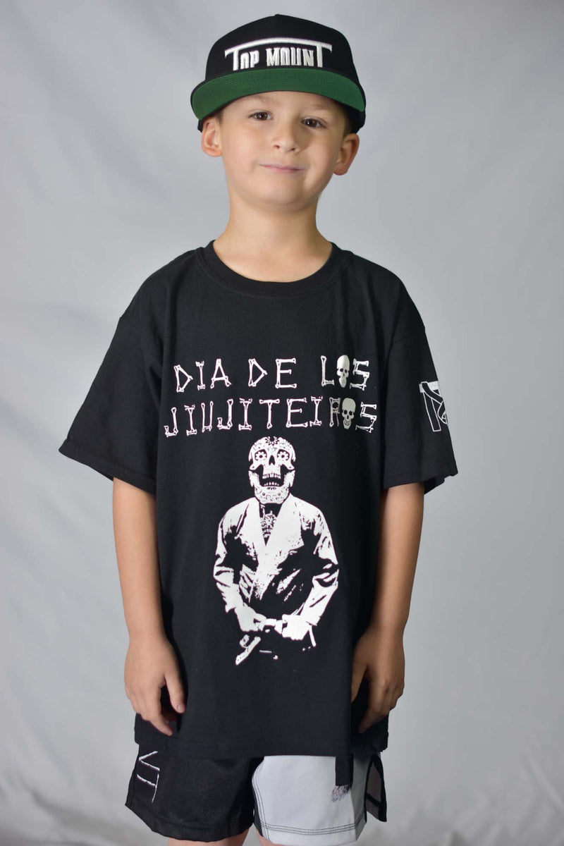 Youth boy in a black Dia De Los Jiujiteiros Jiu Jitsu t-shirt featuring a BJJ sugar skull and Top Mount sleeve logo.