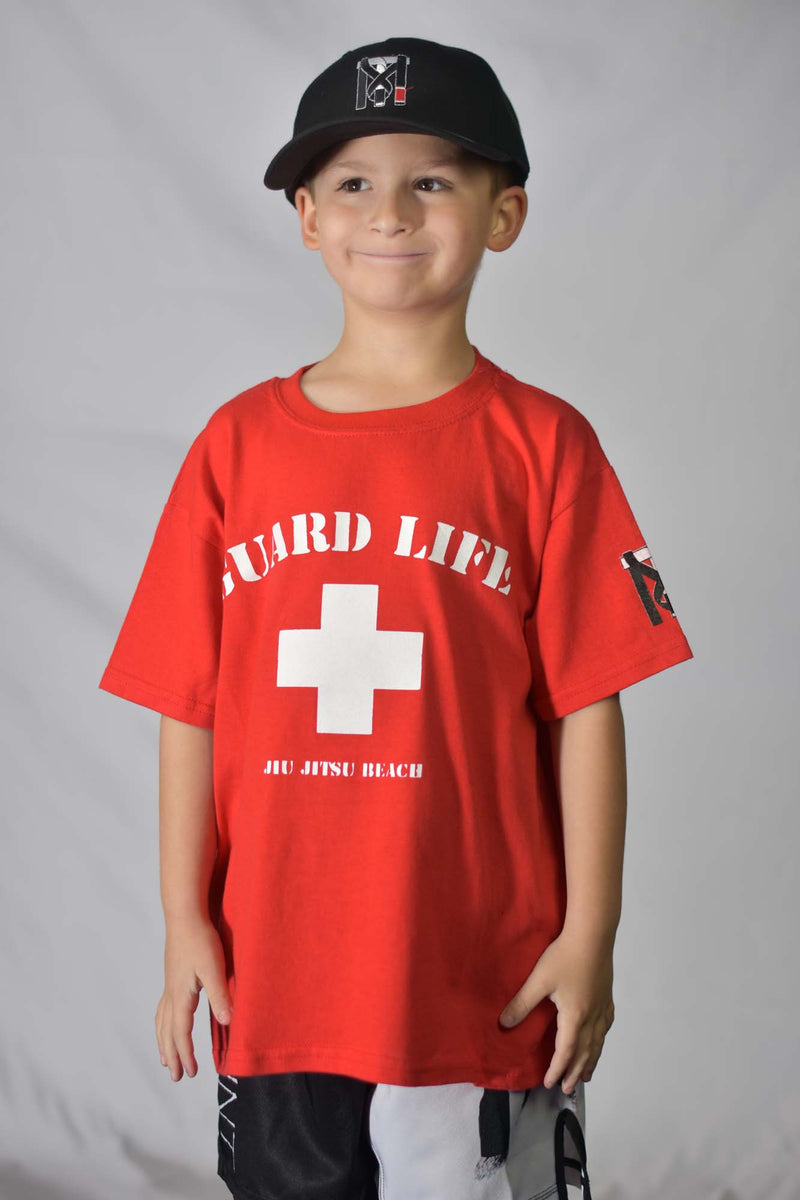 Young boy in red Guard Life Jiu Jitsu Beach graphic tee and black hat. Kids BJJ lifestyle apparel for martial arts.