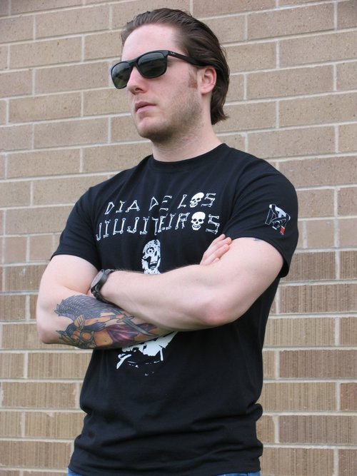 Man in sunglasses wearing a black Dia De Los Jiujiteiros Jiu Jitsu Graphic Tee with sugar skull art and TM sleeve logo.